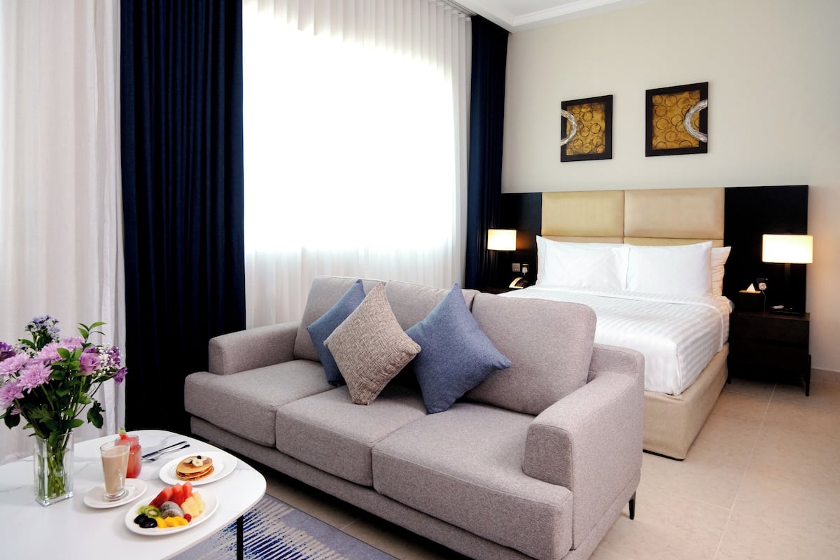 Een hotelkamer met een grijze bank, een bed met witte lakens, een nachtkastje met een lamp en een tafel met ontbijtartikelen.