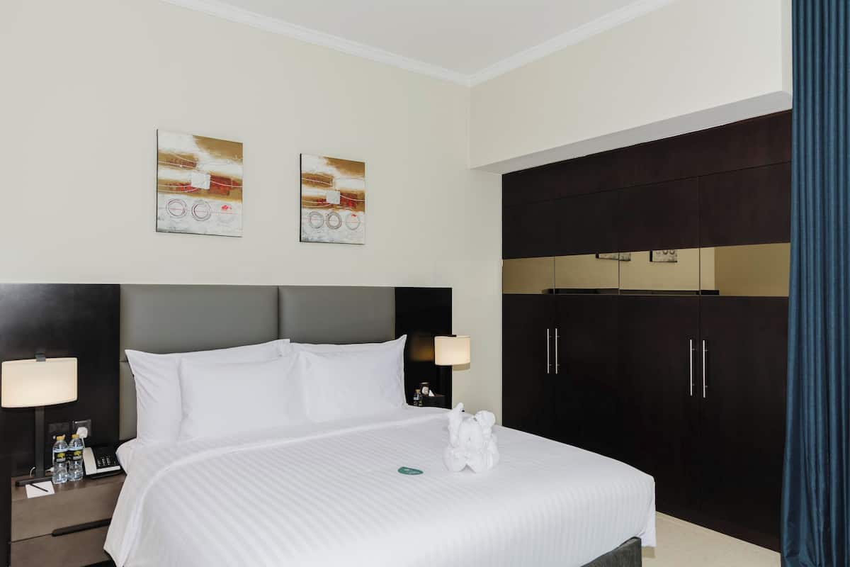Een hotelkamer met een groot bed, twee aan de muur hangende schilderijen, een donkere garderobe en een nachtkastje met een lamp en waterflessen.