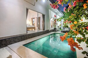 Una piscina con borde de piedra, un dormitorio con cama y cortinas, y vistas al exterior.