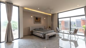 A modern bedroom with a large bed, a small table, and a chair. There is a painting on the wall and a balcony with a view of buildings.