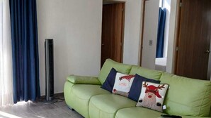 Un sofá verde con almohadones decorativos, un ventilador de pie y una ventana con cortinas azules.