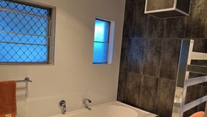 Une salle de bain moderne avec une baignoire blanche, une fenêtre ornée d’une grille métallique et une petite fenêtre située au-dessus de la baignoire.