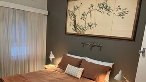 Un lit bien fait, avec du linge de lit marron, deux lampes de chevet, une étagère fixée au mur et une grande œuvre d’art encadrée au-dessus du lit.