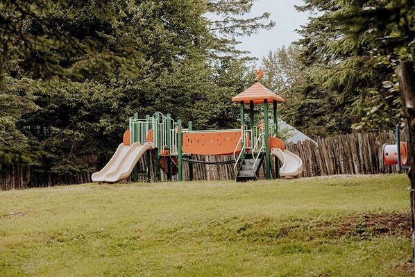 Un parque infantil con toboganes y estructuras para escalar, rodeado de árboles y una valla de madera.
