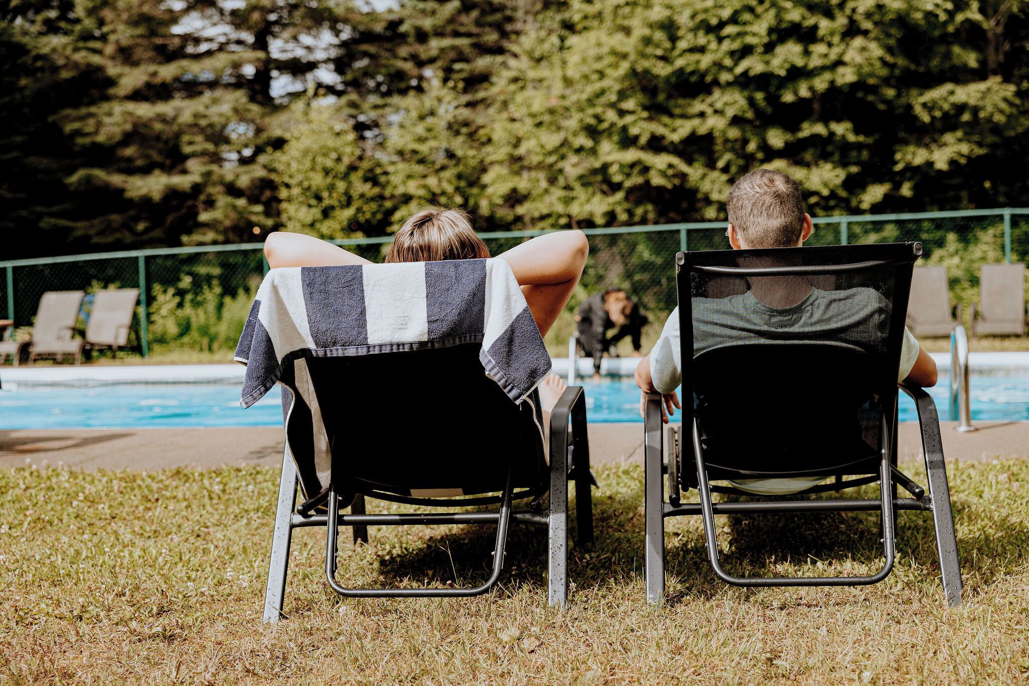Two people sitting on lawn chairs by a pool, with a green fence and trees in the background.