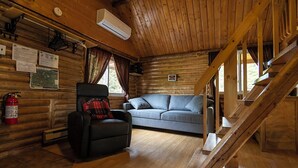 Interior de una cabaña de troncos con sofá, sillón, suelo de madera, escalera y extintor.