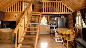 Interior de una cabaña con escalera de madera, cocina, comedor y un dormitorio superior con una colcha roja.