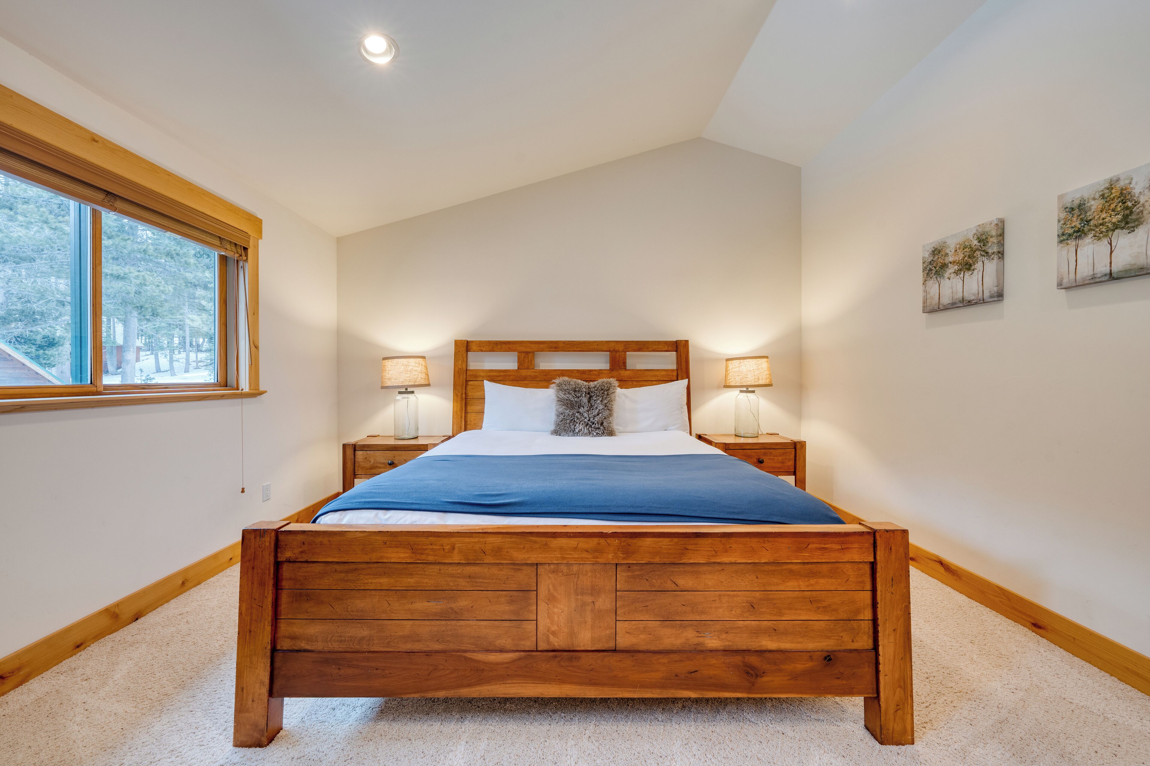 Une chambre à coucher avec un lit en bois, deux lampes de chevet, une fenêtre donnant sur des arbres et deux tableaux accrochés au mur.