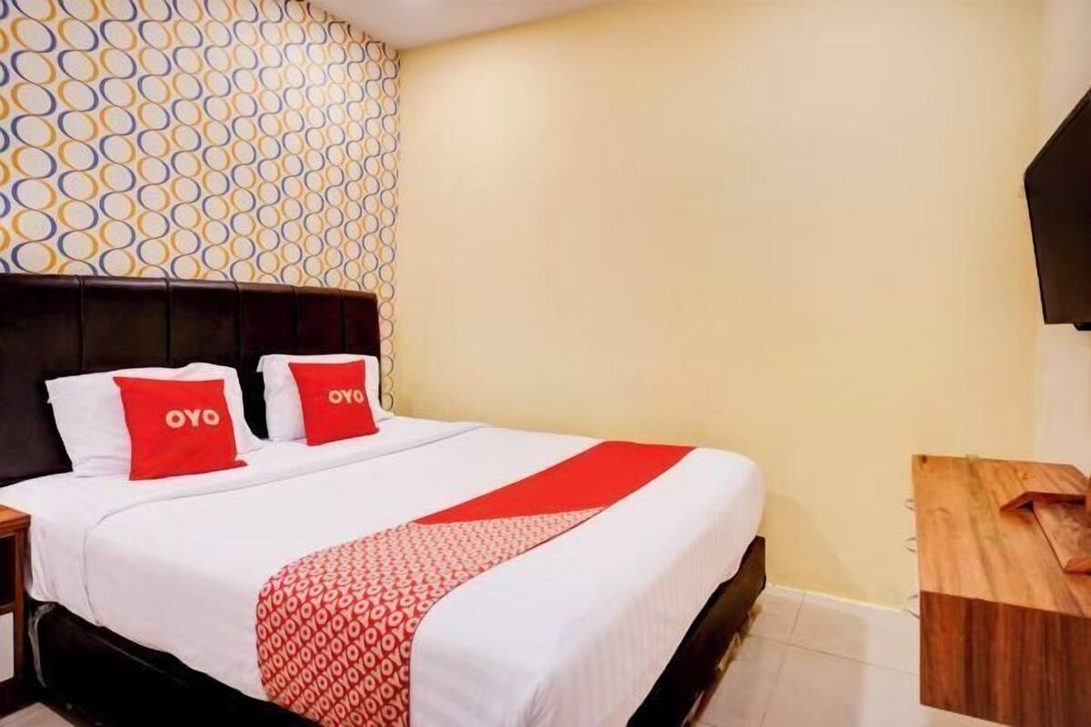 Une chambre d’hôtel comprenant un lit, deux oreillers à l’effigie de la marque « OYO », un mur décoré d’un motif, une table de chevet en bois et un bureau en bois.