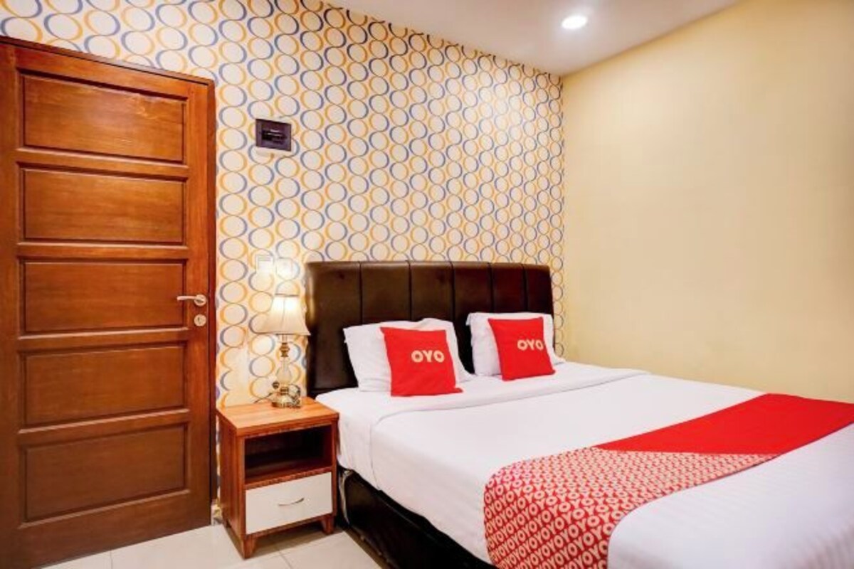 A hotel room with a patterned wallpaper, a wooden door, a bedside table with a lamp, a bed with white bedding and red pillows, and a red throw blanket.
