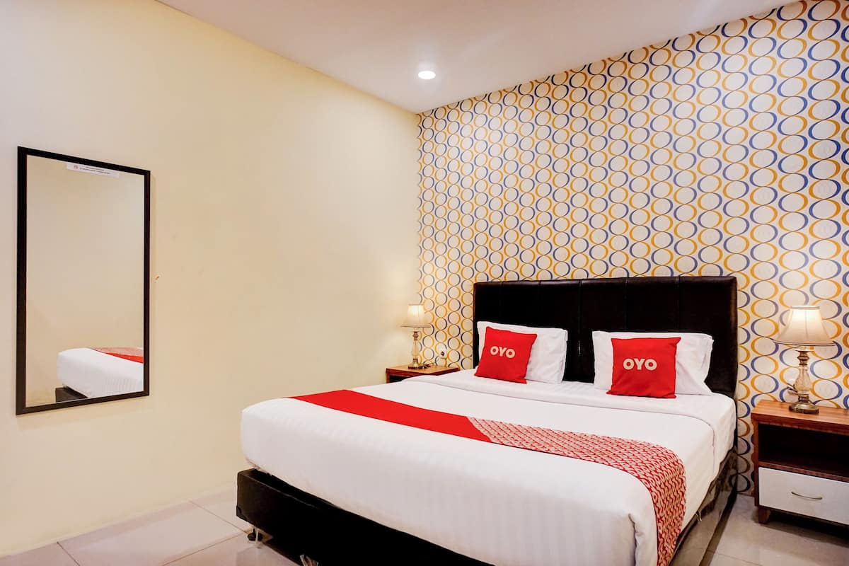 A neatly made bed with a red and white bedspread, two red pillows with "OYO" branding, a bedside table with a lamp, and a patterned wallpaper.
