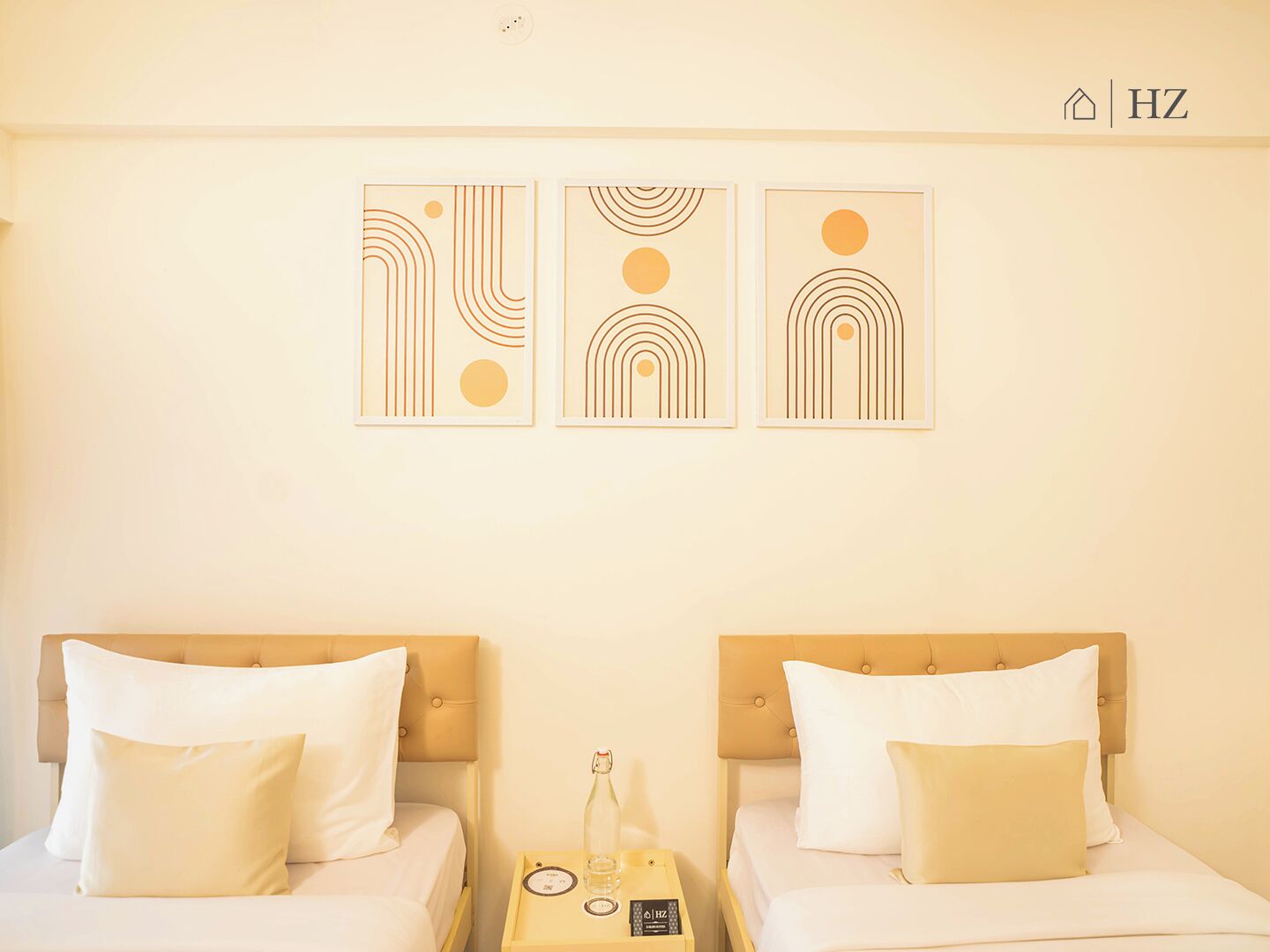 Hotellihuone, jossa on kaksi sänkyä, ja jokaisessa sängyssä on pääty sekä yöpöytä, jossa on pullo ja kaukosäädin. Lisäksi huoneessa on kolme kehystettyä, abstraktia seinätaideteosta.