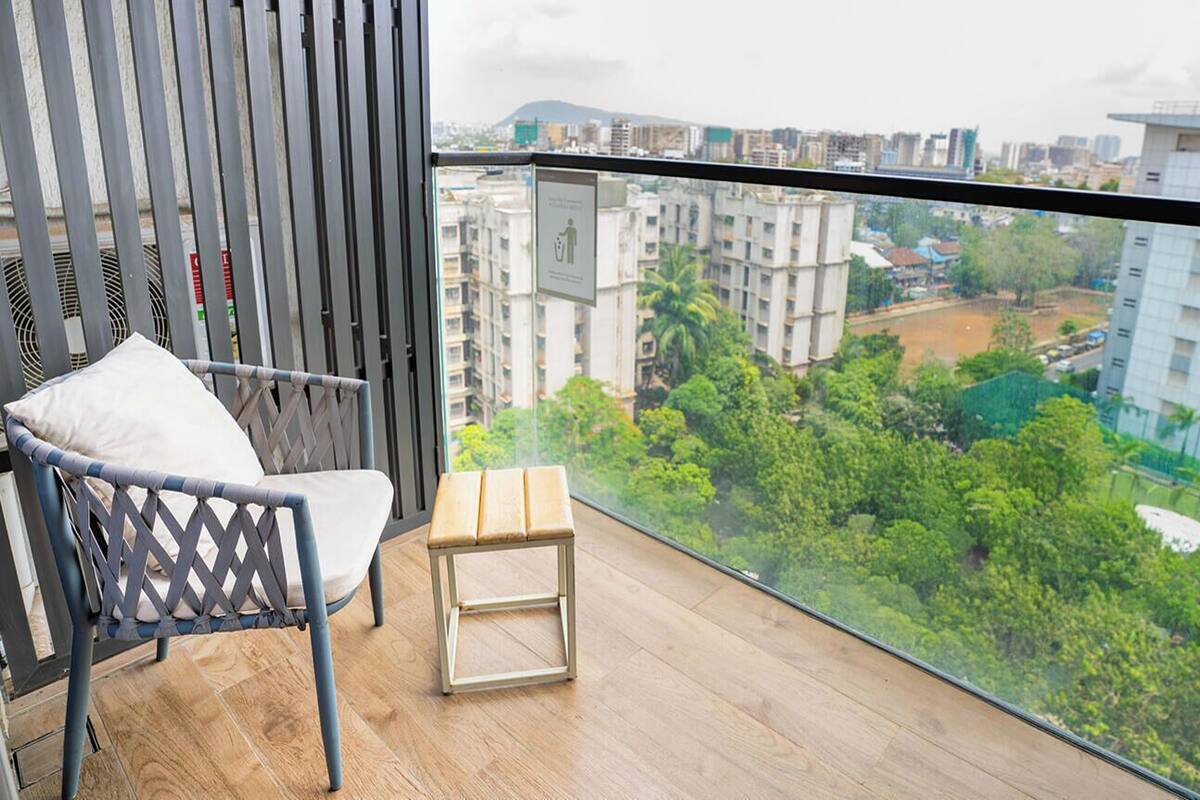A balcony with a chair and a small table, overlooking a cityscape with buildings and greenery.