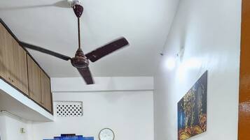 A room with a ceiling fan, a wooden cabinet, a white sofa, green chairs, a clock, and a wall with a picture.