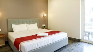 A neatly made bed with white and red linens, a headboard, and a small bedside table with a lamp. - Arooba Residency Calangute (Calangute)