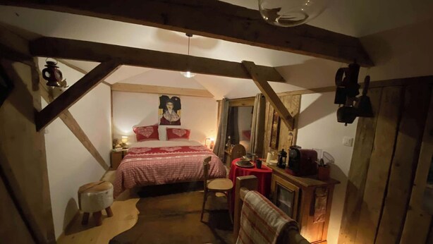 Une chambre mansardée chaleureuse, avec un plafond en pente, un lit avec une literie rouge, une chaise en bois, une petite table recouverte d’une nappe rouge et un tapis en peau de vache sur le sol.