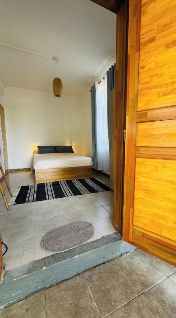 Un dormitorio con cama, una puerta de madera, una ventana con cortinas, una alfombra pequeña y una lámpara fijada a la pared.