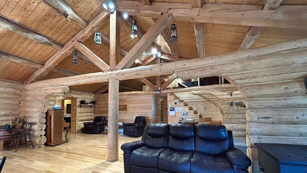 Interior de una cabaña de troncos con piso de madera, sofá de cuero negro y una escalera que sube a un nivel superior.