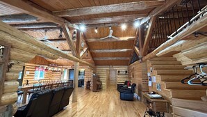 Interior de una cabaña de troncos con piso de madera, un sofá, una mesa de comedor, una escalera y una lámpara colgante.