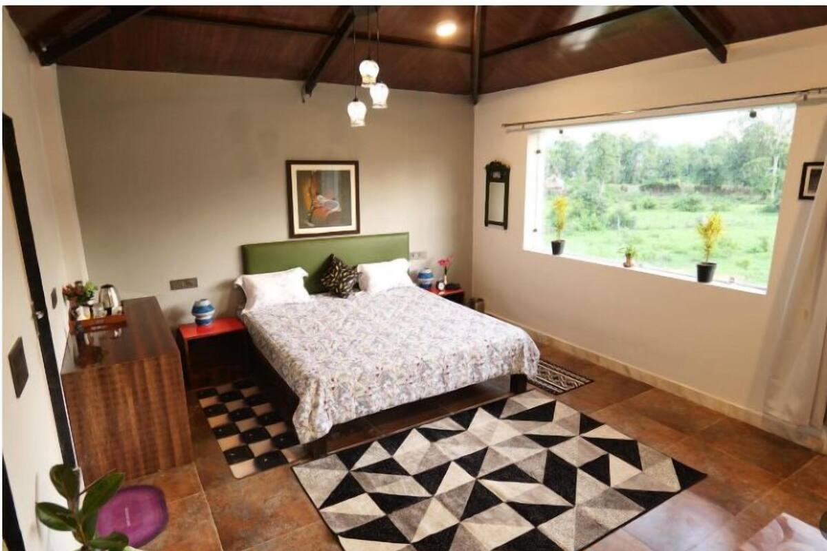 Une chambre à coucher comprenant un lit, une table de chevet, une commode, une fenêtre donnant sur un paysage verdoyant et un tapis à motifs géométriques.