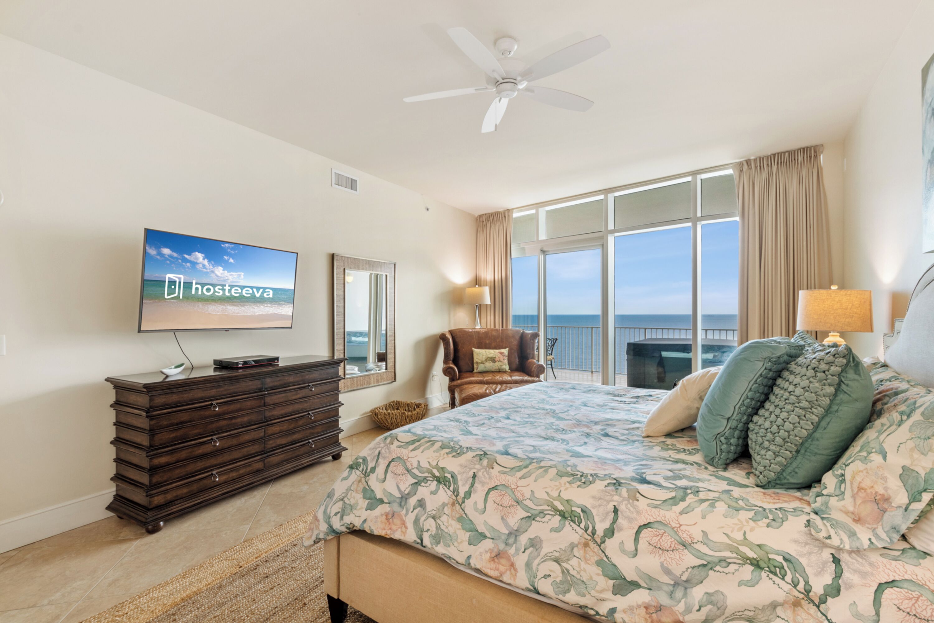 A bedroom with a large bed, a television on the wall, a ceiling fan, a view of the ocean, and a window with curtains.