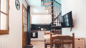 A kitchen with white cabinets, a wooden dining table with chairs, a black TV mounted on the wall, and a wooden staircase. - Le Colimaçon - Half-timbered house (Tours)