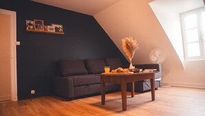 A cozy living room with a dark blue wall, a wooden floor, a grey sofa, a small table with a vase of dried flowers, and a window with natural light.