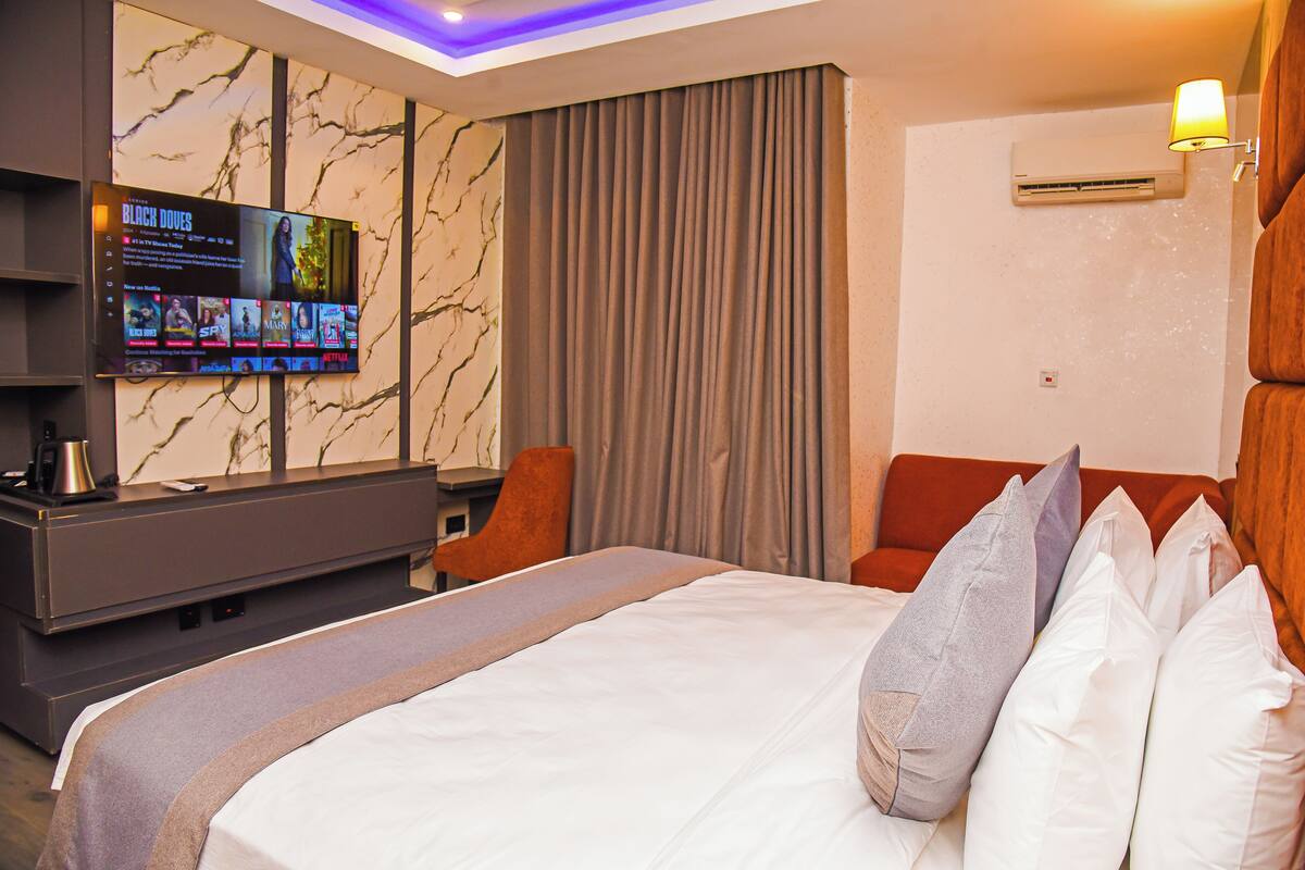 A modern hotel room with a large bed, a flat-screen TV mounted on the wall, a desk with a chair, and a window with curtains.