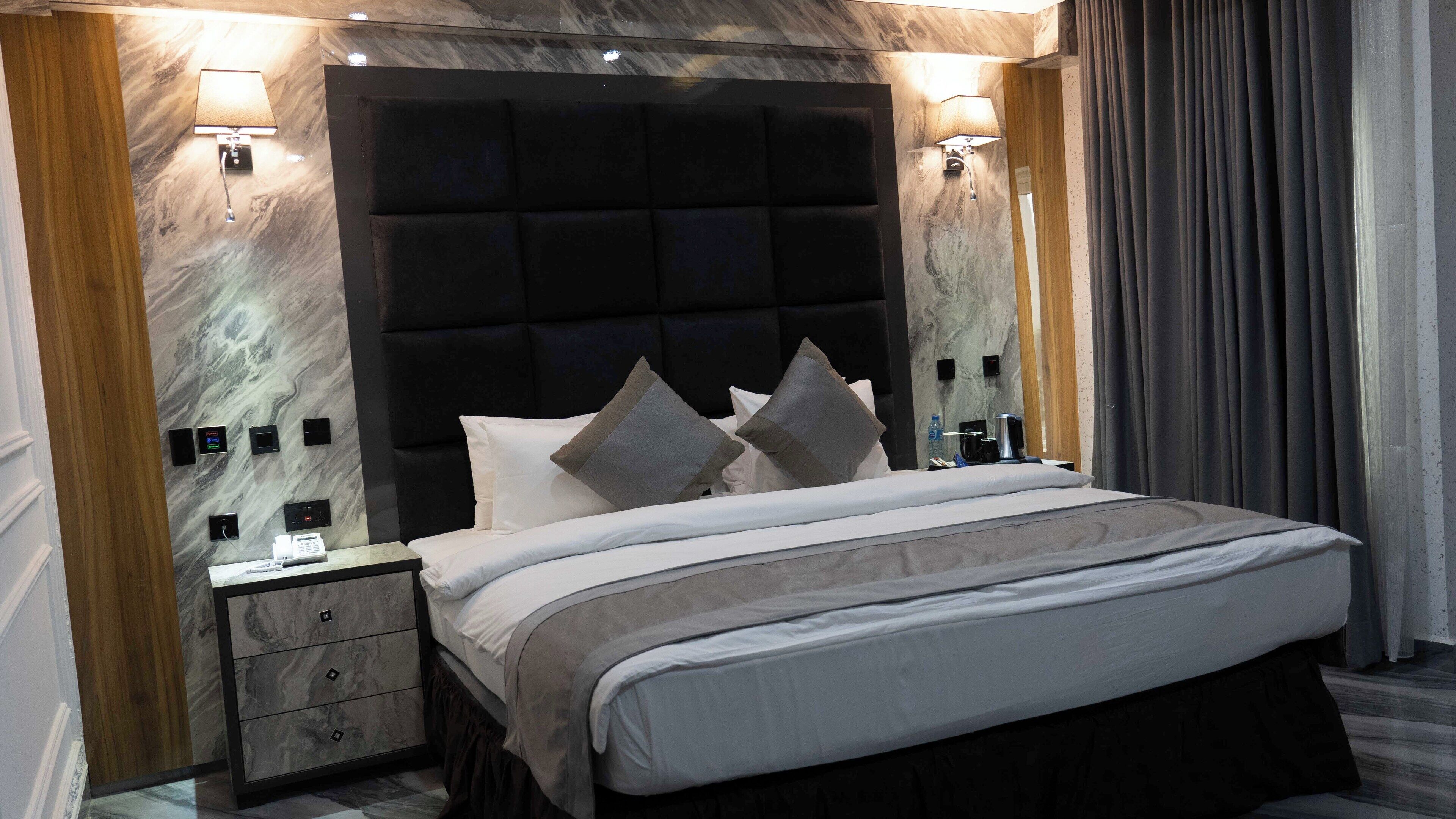 A modern hotel room with a large bed, a wooden nightstand, a marble wall, and a dark headboard.