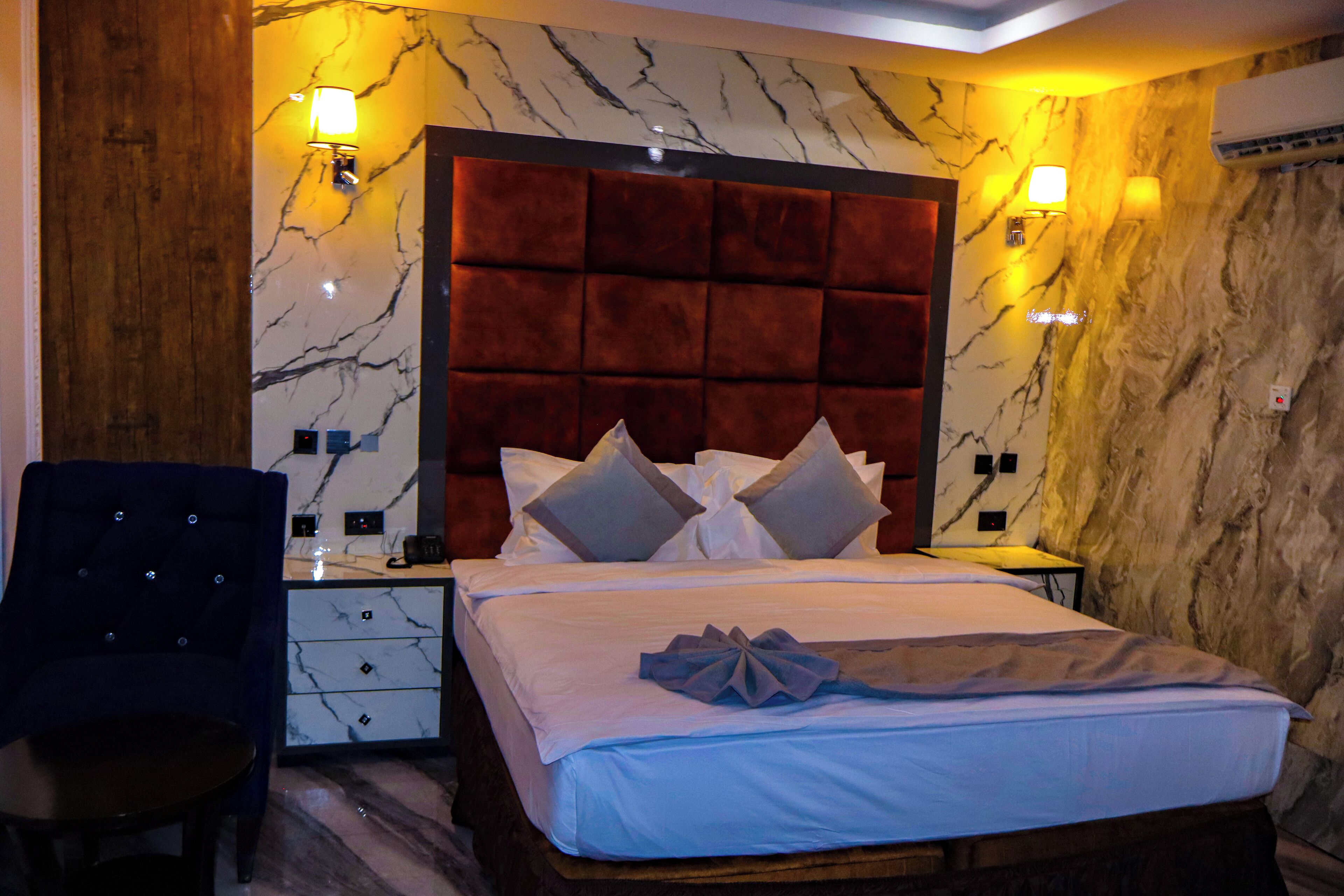 A hotel room with a marble-patterned wall, a tufted headboard, a bed with pillows, a nightstand, a chair, and a table.