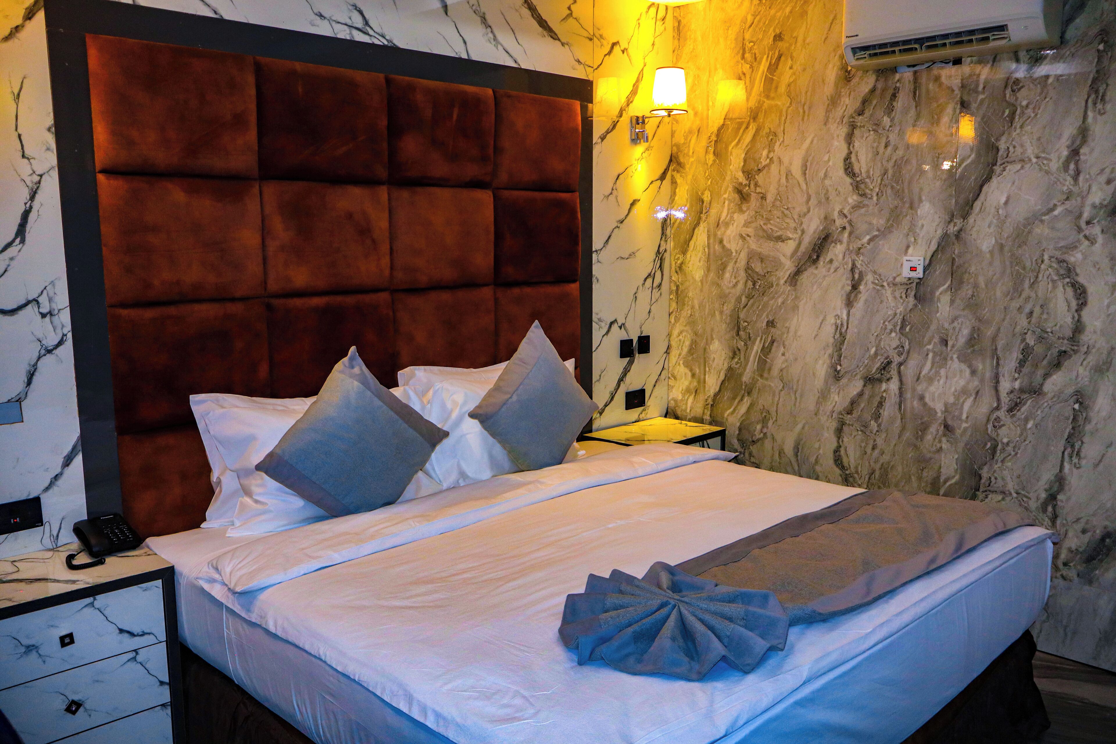 Une chambre d’hôtel avec une tête de lit capitonnée, un mur en marbre, un lit avec du linge de lit blanc et des oreillers, une table de chevet avec une lampe et un appareil de climatisation.