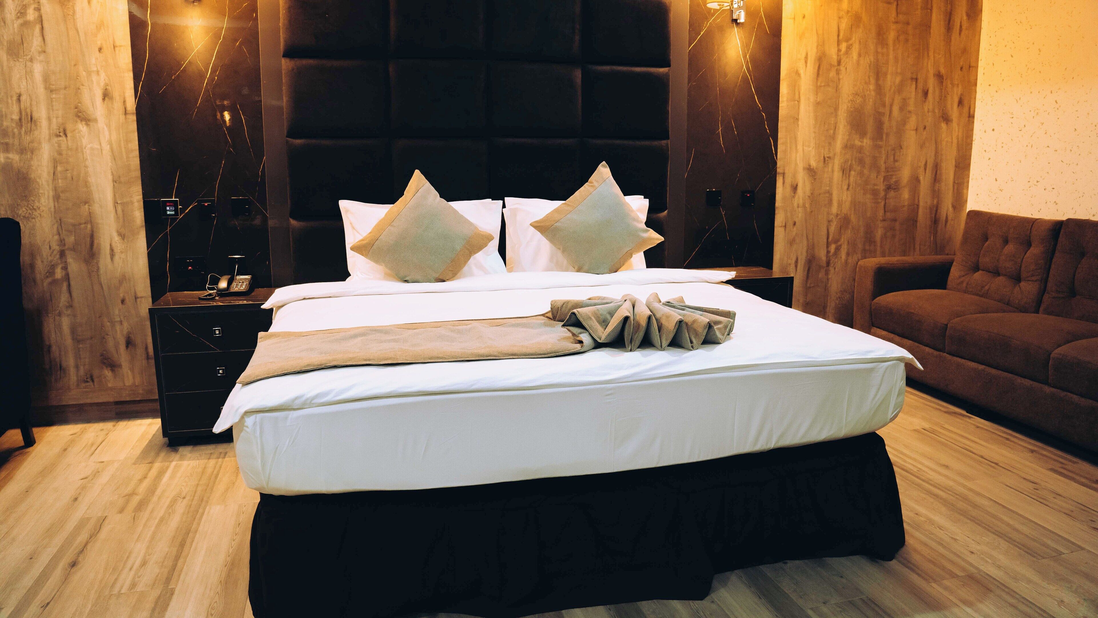 A neatly made bed with white linens and multiple pillows, a wooden nightstand with a phone and a lamp, and a brown leather sofa in a modern room with wooden walls and a large padded headboard.