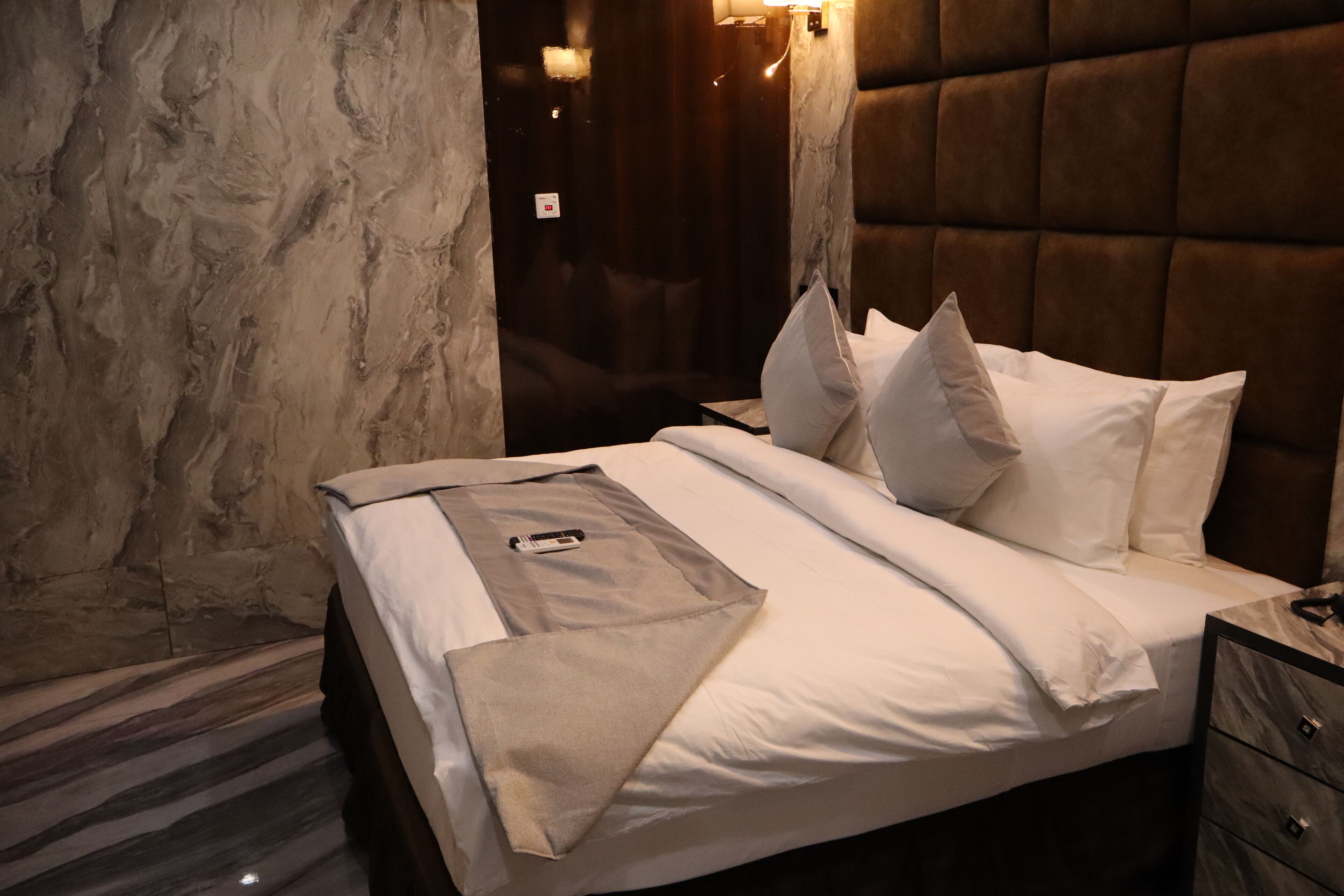 Una cama bien tendida con sábanas y almohadas blancas, una mesita de noche con lámpara y una pared con diseño de mármol.