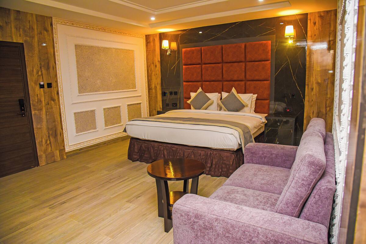 A hotel room with a large bed, a purple sofa, a small table, and a wooden floor.