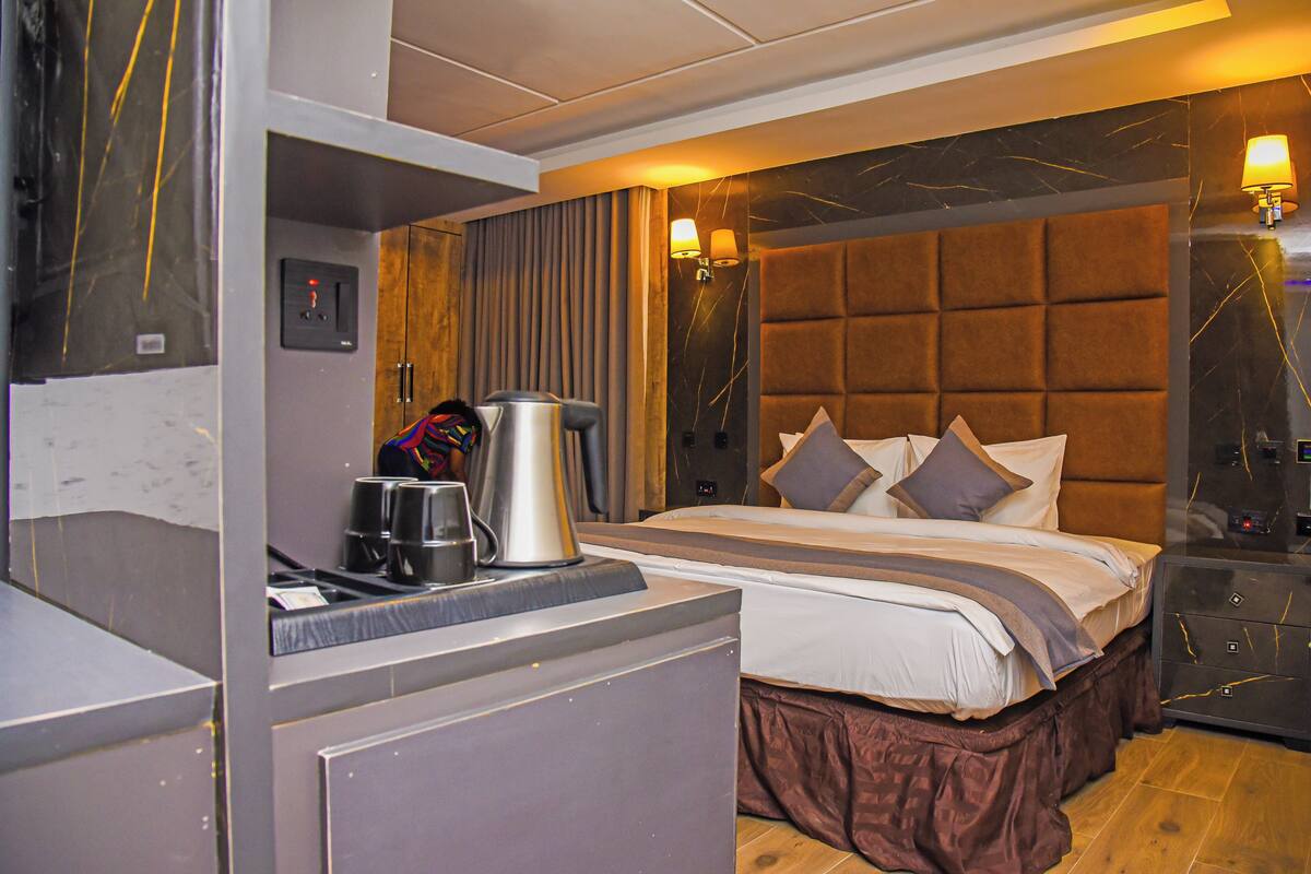 A modern hotel room with a large bed, a bedside table with a kettle, and a nightstand with a lamp.