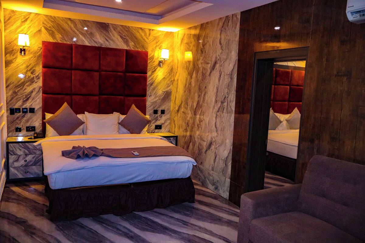 A hotel room with a large bed, marble walls, and a red headboard. There is a nightstand with a lamp and a chair in the corner.