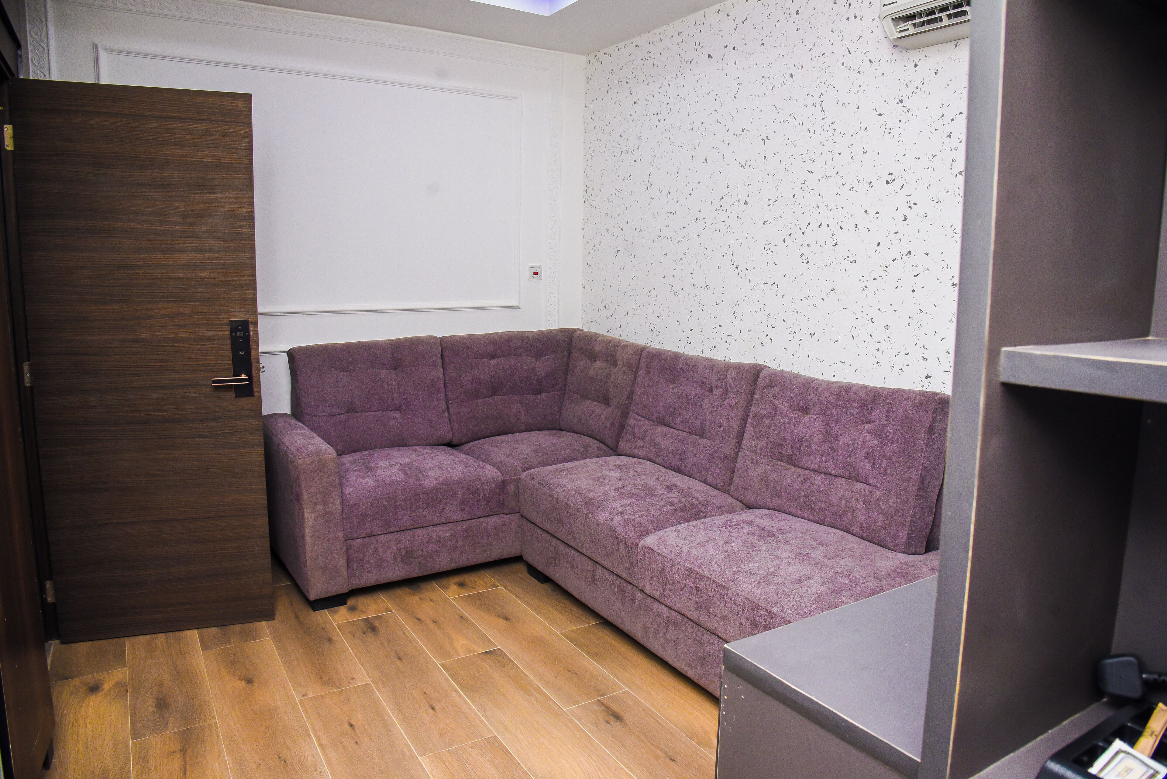 A corner room with a purple sectional sofa, wooden flooring, a white door, and a wall-mounted air conditioning unit.