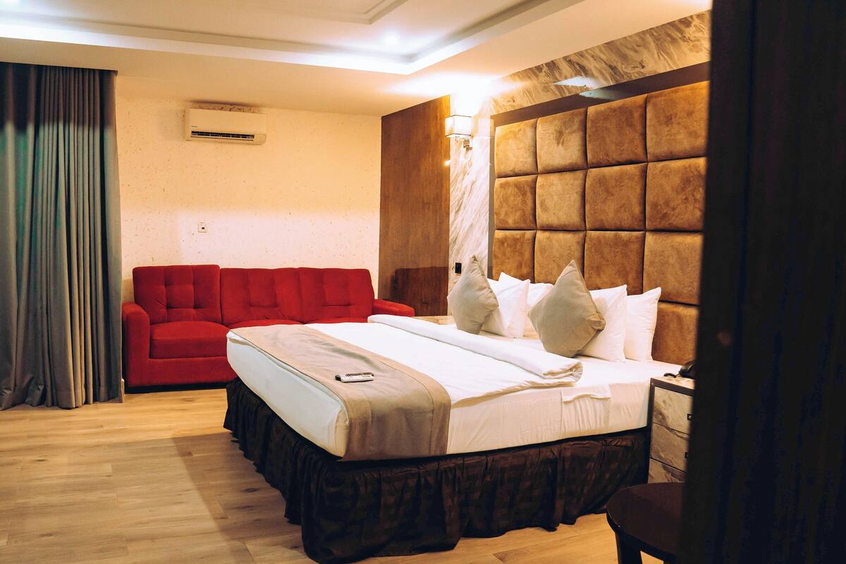 A hotel room with a bed, a red sofa, a wooden floor, and a window with curtains.