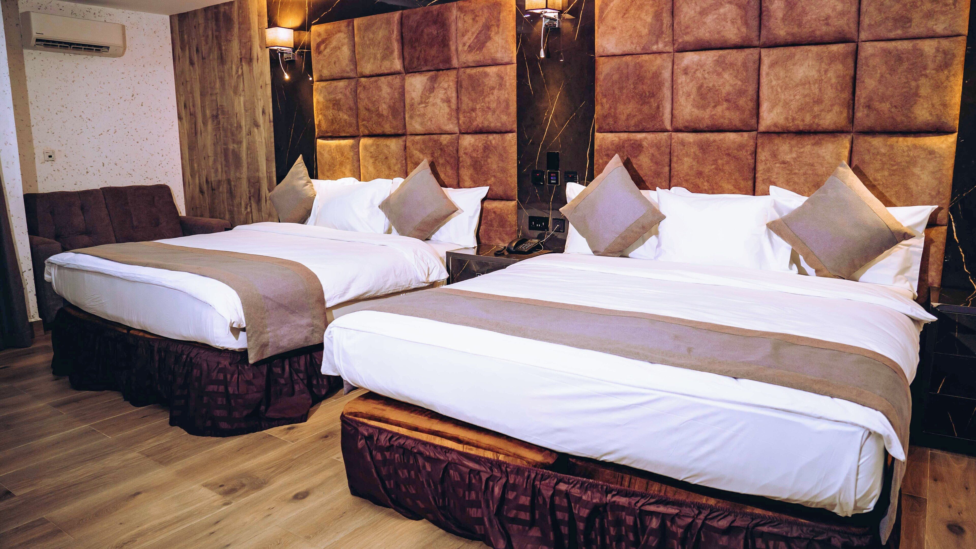 A hotel room with two beds, wooden flooring, a large padded headboard, and a wall-mounted air conditioning unit.