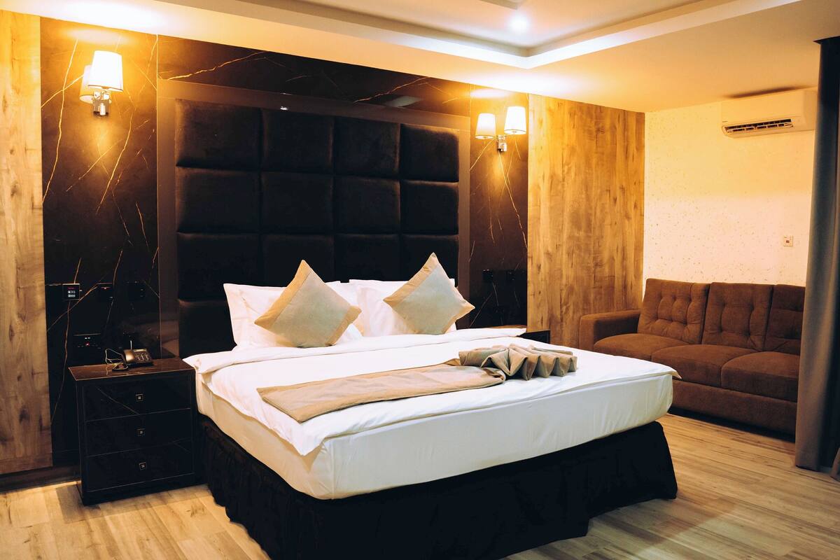 A hotel room with a large bed, a wooden headboard, two bedside lamps, a brown sofa, and a curtain.