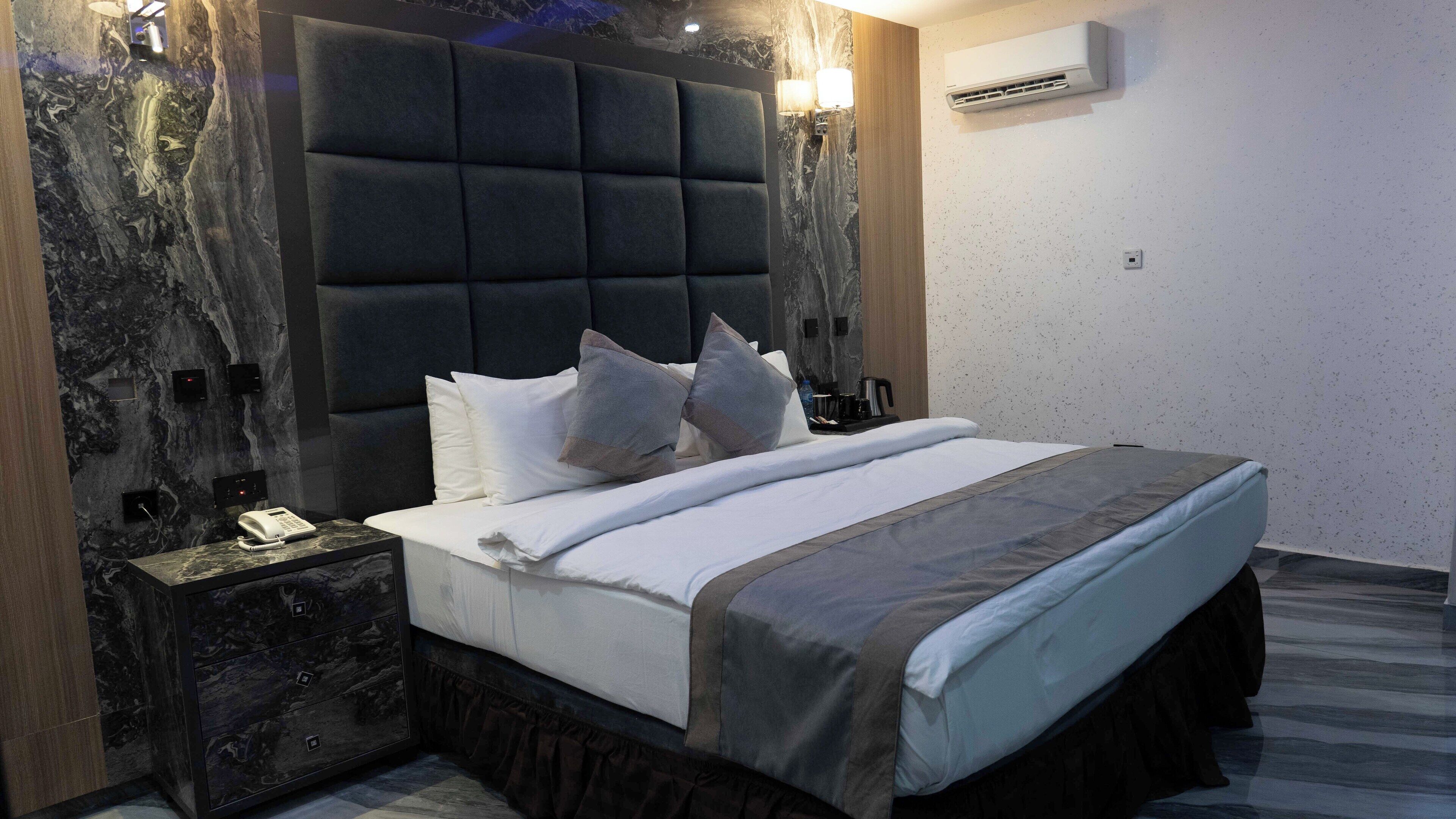 A modern hotel room with a marble wall, a large bed with a padded headboard, a bedside table with a phone, and an air conditioning unit on the wall.