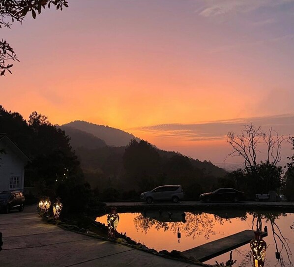 Una puesta de sol serena sobre una cadena montañosa, una masa de agua tranquila reflejando el cielo y un estacionamiento con coches y una casa.