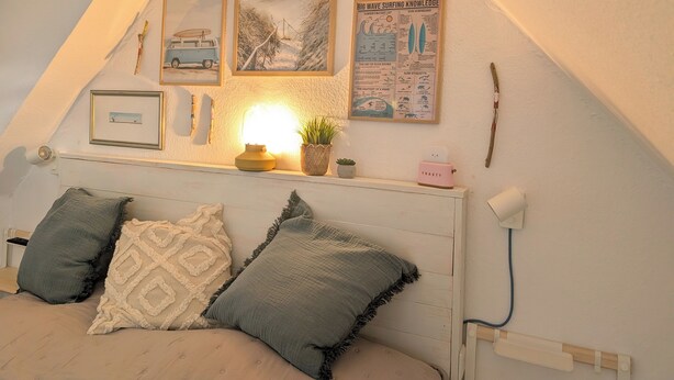 A cozy attic bedroom with a sloped ceiling, a bed with two pillows, a bedside lamp, and a shelf with a plant and a framed surfboard. - Haus Hanne Sylt ♥ Apartment OG- Maisonette in Alt Westerland. Families welcome! (Sylt)