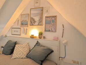 Ein gemütliches Schlafzimmer im Dachgeschoss mit schrägen Wänden, einem Bett mit zwei Kissen, einer Nachttischlampe und einem Regal mit Pflanze und gerahmtem Surfbrett.