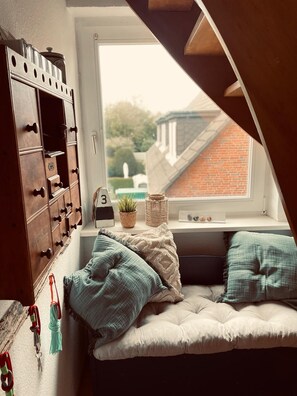 Een knus zolderkamer met een inbouwed bed, een houten opbergunit en een raam met uitzicht op een rijtjeshuis.
