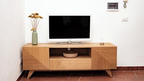 Ett modernt vardagsrum med en platt-TV placerad på en träkonsol, en vas med torkade blommor och en väggmonterad telefon.