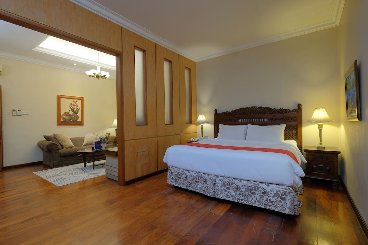Quarto de hotel com uma cama grande, área de estar com sofá e mesa de centro, painéis de madeira e um candelabro.