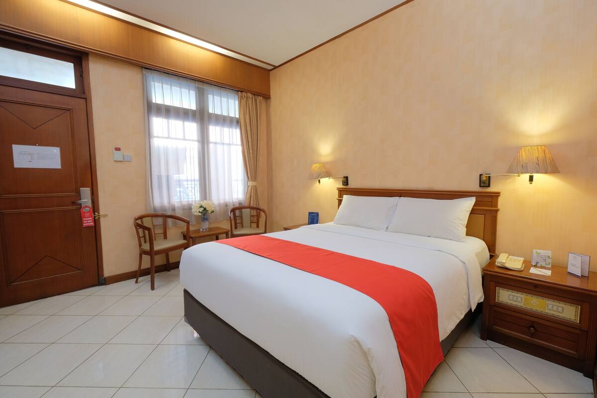 Quarto de hotel com uma cama grande, mesa de cabeceira de madeira, uma cadeira, escrivaninha e janela com cortinas.