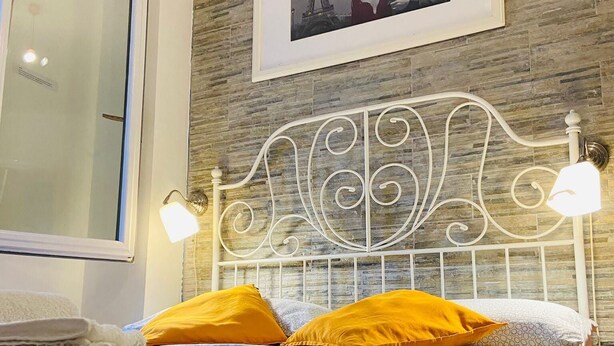 A neatly made bed with patterned bedding and two yellow pillows, a white metal headboard, and a framed picture of the Eiffel Tower on the wall. - Europe station rooms (Parma)