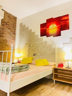 A bedroom with a white metal bed frame, a brick wall, a staircase, and a sunset painting on the wall. - Europe station rooms (Parma)